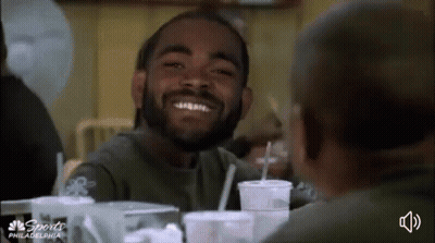 Bearded Man Smiling At Another At Table With Drinks GIF