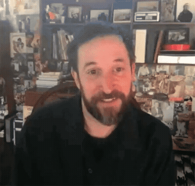 Bearded Man Smiling In Front Of Bookshelf With Pictures GIF