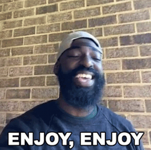 Bearded Man Smiling In Front Of Brick Wall Saying Enjoy Enjoy GIF