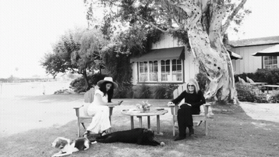 Black And White Photo Of Two Women With Dogs In Front Of A House GIF