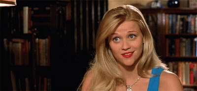 Blonde Woman In Blue Top Smiling In Front Of Bookshelf GIF