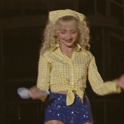 Blonde Woman In Yellow Shirt And Blue Shorts Holding Microphone GIF