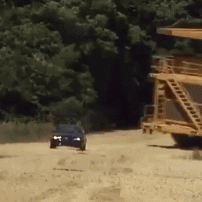Blue Car Driving Down Dirt Road Past Yellow Truck GIF