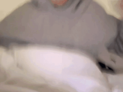 Blurry Person Laying On Bed In Gray Sweater GIF