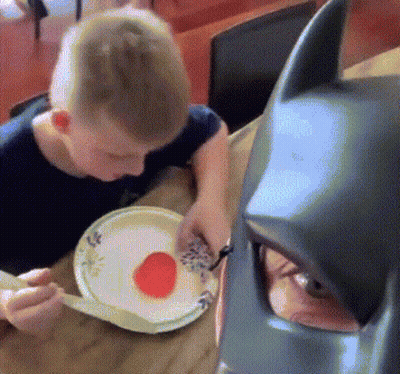 Boy Eating Plate Of Food Next To Batman Mask GIF
