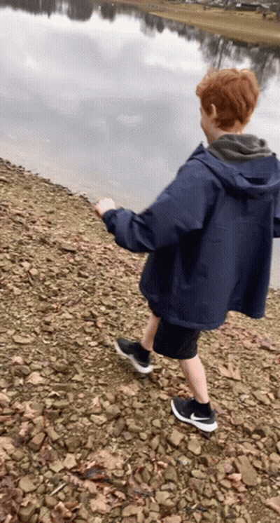 Boy In Blue Jacket Walking On Rocky Shoreline GIF