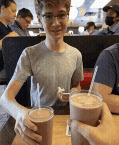 Boy With Glasses Sitting At Table With Two Milkshakes GIF