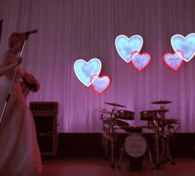 Bride Singing Into Microphone In Front Of Drum Set With Hearts GIF