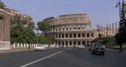 Cars Are Driving Down A Street In Front Of A Large Building Called The Colosseum GIF