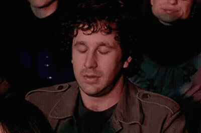 Curly-Haired Man In Crowd Eyes Closed Hand On Chin GIF
