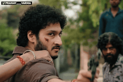 Curly Haired Man With Beard Holding Gun In Front Of Pranay Varma Sign GIF