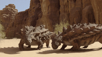 Dinosaurs Standing In Sand Near Rock Formation GIF
