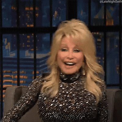 Dolly Parton Smiling In Chair In Front Of City Skyline GIF