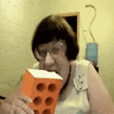 Elderly Woman Holding Brick In Mouth With Glasses GIF
