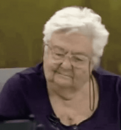 Elderly Woman In Glasses And Purple Shirt Smiling On Couch GIF