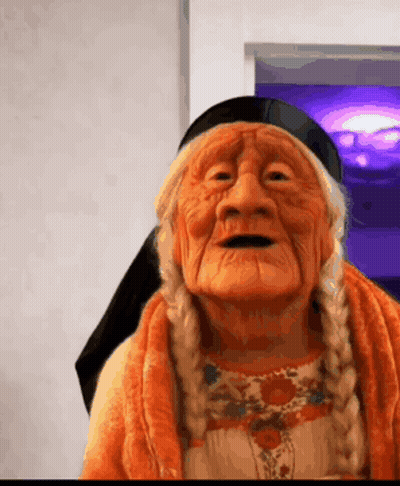 Elderly Woman With Braids In Orange Sweater And Black Scarf GIF