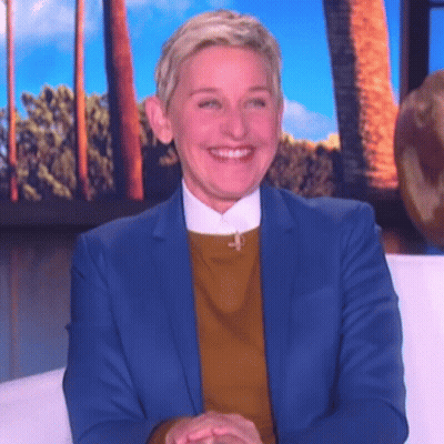 Ellen Degeneres Is Wearing A Blue Suit And A Yellow Sweater And Smiling While Sitting On A Couch . GIF