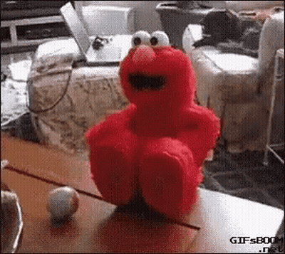 Elmo The Sesame Street Character Is Sitting On A Table With A Ball In Front Of Him . GIF