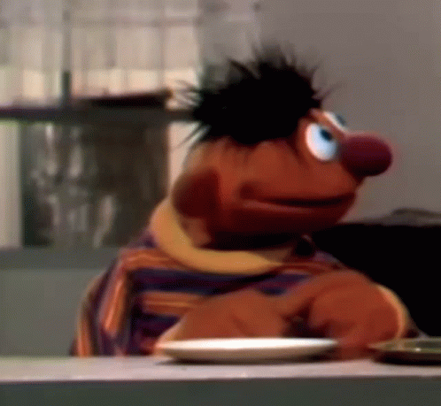 Ernie From Sesame Street Is Sitting At A Table With A Plate Of Food In Front Of Him . GIF