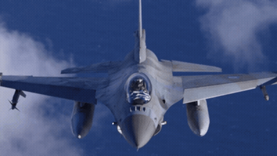 Fighter Jet Flying Through Cloudy Blue Sky With Flag In Cockpit GIF