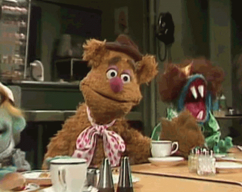 Fozzie Bear Is Sitting At A Table With A Cup Of Coffee GIF