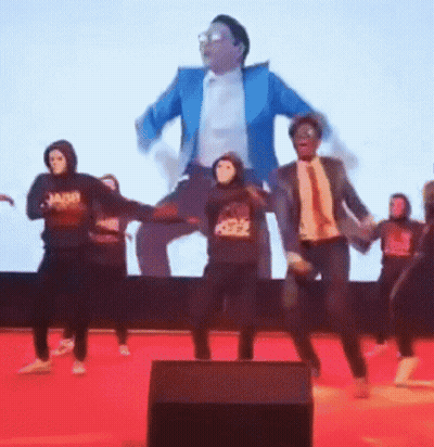Group Dancing In Masks In Front Of Tuxedo Man On Screen GIF