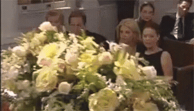 Group Sitting In Front Of Coffin Covered In Flowers GIF