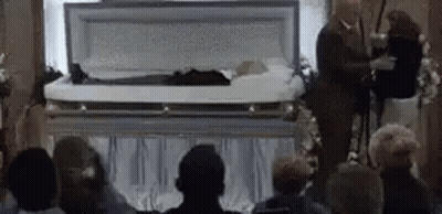 Group Sitting In Front Of Coffin With Man Standing GIF