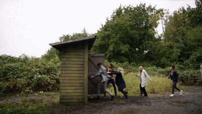 Group Standing Around Wooden Outhouse GIF