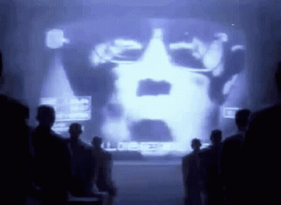 Group Standing Before Large Screen Showing Man's Face GIF