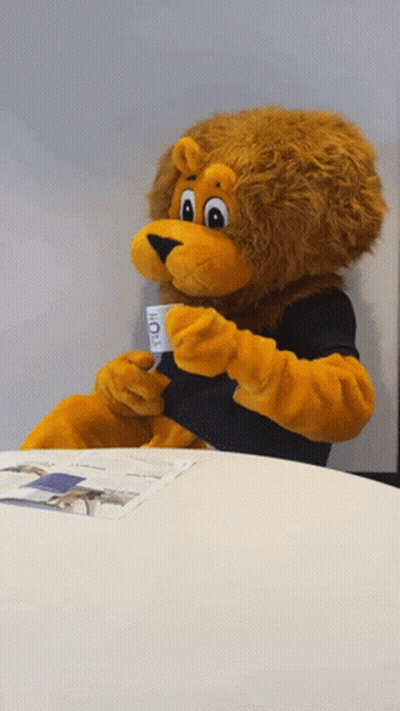 Lion Mascot Sitting At Table Holding Coffee Cup GIF