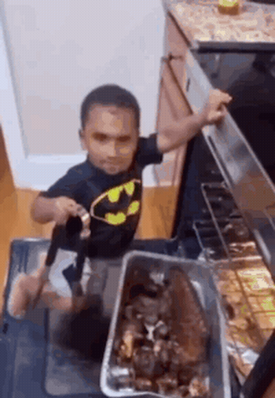 Little Boy In Batman Shirt Standing By Oven With Pan Of Food GIF