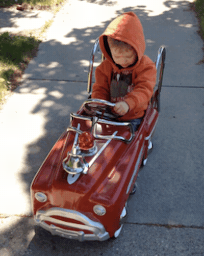 Little Boy In Orange Hoodie Sitting In Red Toy Car GIF