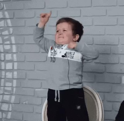 Little Boy In World Coaster Sweatshirt In Front Of Brick Wall GIF
