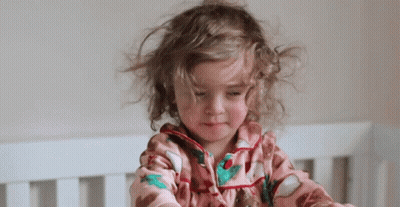Little Girl With Curly Hair Sitting In Crib Looking At Camera GIF