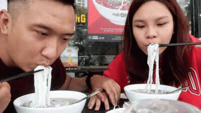 Man And Woman Eating Noodles With Chopsticks In Front Of Ung Sign GIF