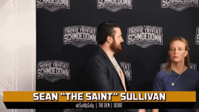 Man And Woman In Front Of Movie Trivia Schmoodown Wall GIF