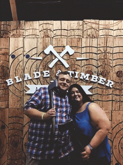 Man And Woman Posing In Front Of Blade And Timber Sign GIF