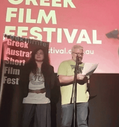 Man And Woman Standing At Greek Film Festival Sign GIF