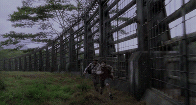 Man And Woman Walking Through Fenced Area With Trees GIF