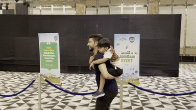 Man Carrying Child In Front Of Welcome To Jorbadar Event Sign GIF