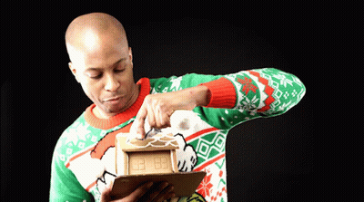 Man Decorating Gingerbread House In Green And Red Sweater GIF