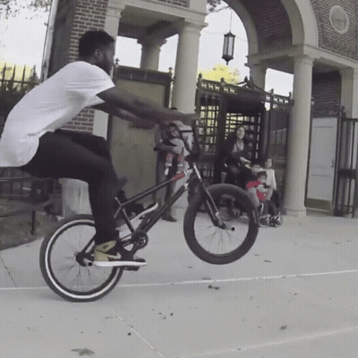 Man Doing Bike Trick In Front Of Brick Building With Sign GIF