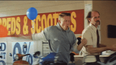 Man Holding Blue Balloon In Front Of Speeds & Scooters Sign GIF