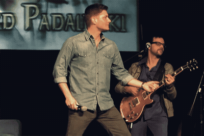 Man Holding Microphone Next To Guitarist In Front Of Padalski Screen GIF