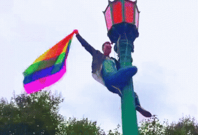 Man Holding Rainbow Flag Hanging From Street Light GIF