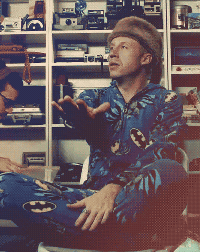 Man In Batman Pajama Sitting In Front Of Camera Shelves GIF