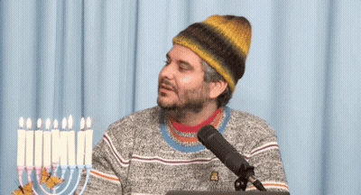 Man In Beanie And Sweater Sitting At Microphone With Menorah GIF