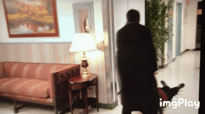 Man In Black Coat In Hallway With Couch And Lamp GIF