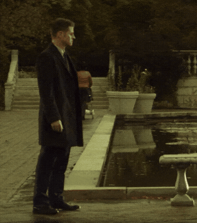 Man In Black Coat Walking By Fountain GIF